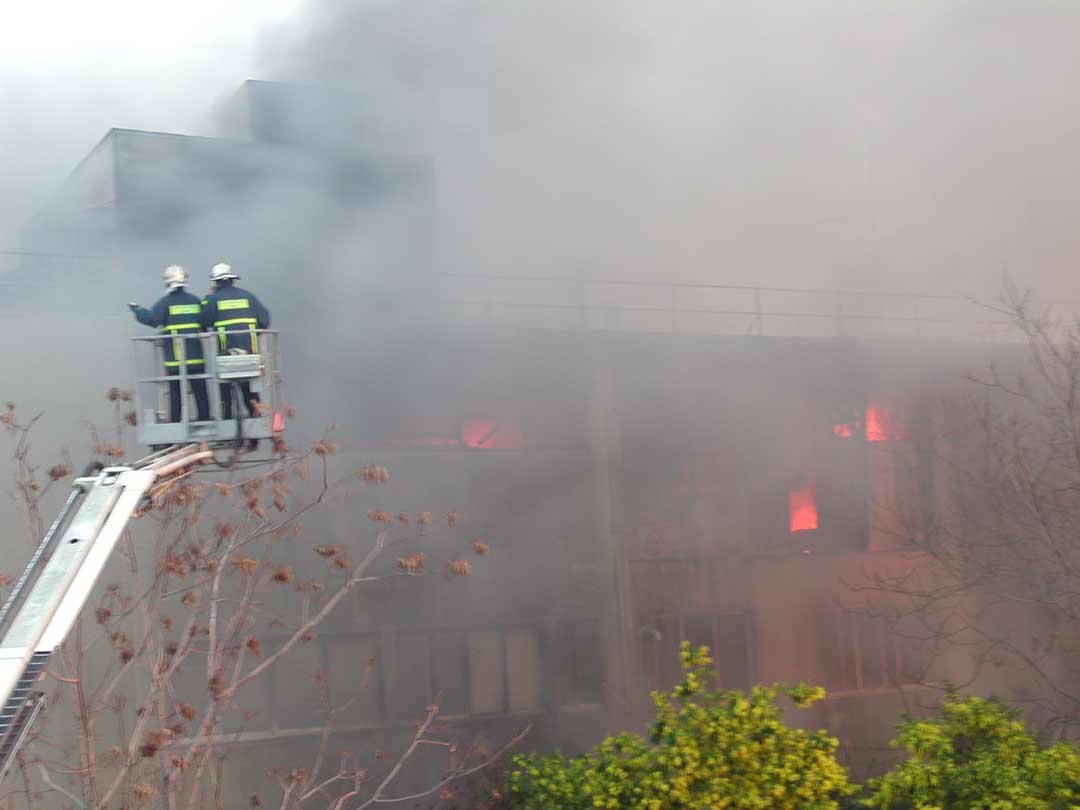 Πυροσβέστες σε καλάθι κλιμακοφόρου οχήματος μπροστά στο εργοστάσιο του Βοτανικού, μέσα σε πυκνό καπνό και φλόγες στα παράθυρα.